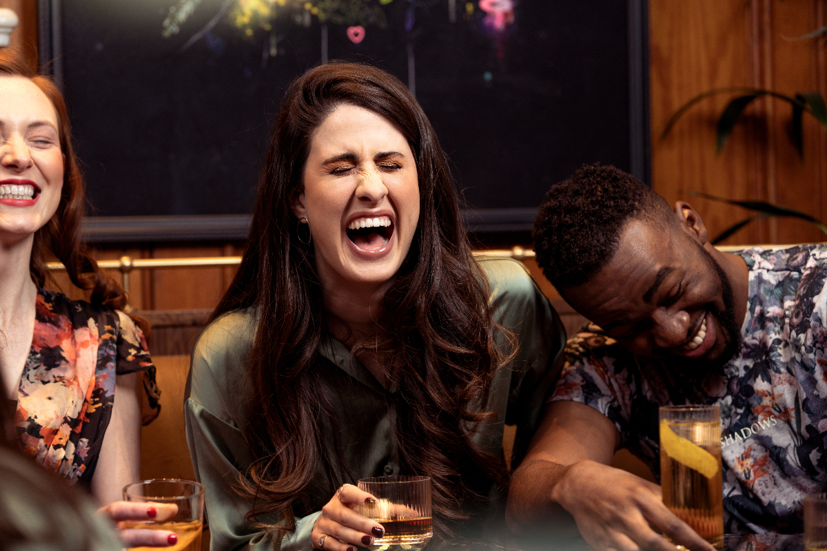 A woman with long brown hair laughs as she sits next to two other friends in a restaurant