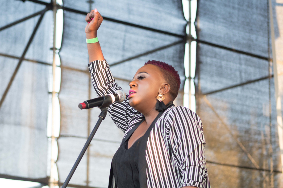 A Black woman with short hair wearing a black-and-white striped top raises her right arm in the air triumphantly.