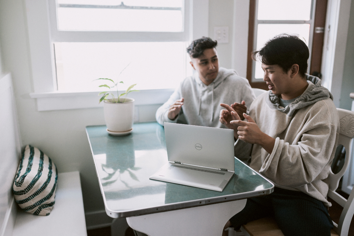 Two men sit side by side at a table discussing something on their Dell laptop.