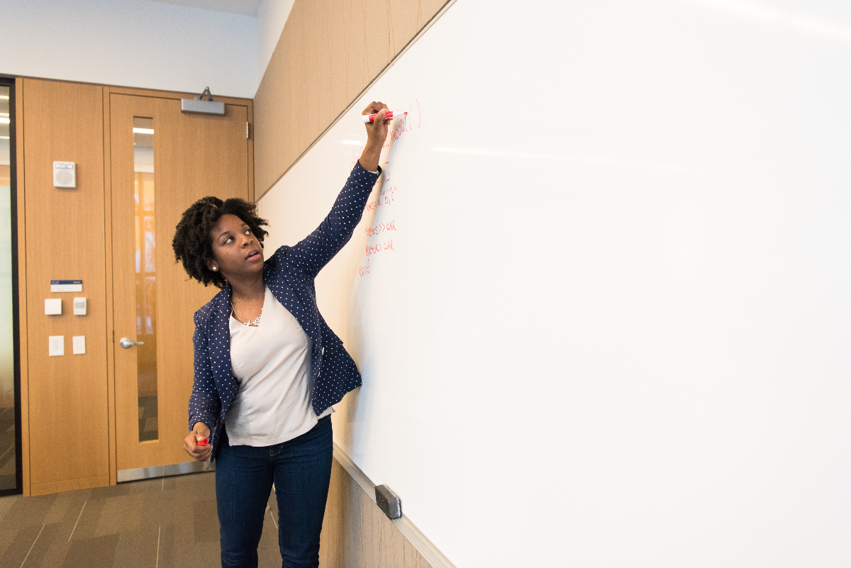 A Black woman in a white top underneath a dark blazer uses a whiteboard to brainstorm for Cisco interview questions.