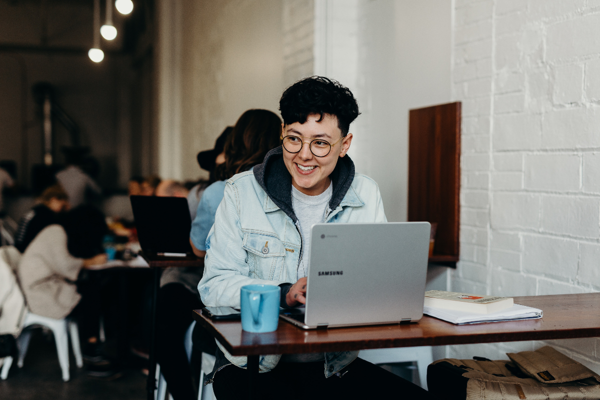 A person with short, dark brown hair smiles while they work on learning some high income skills on their laptop in a coffee shop.