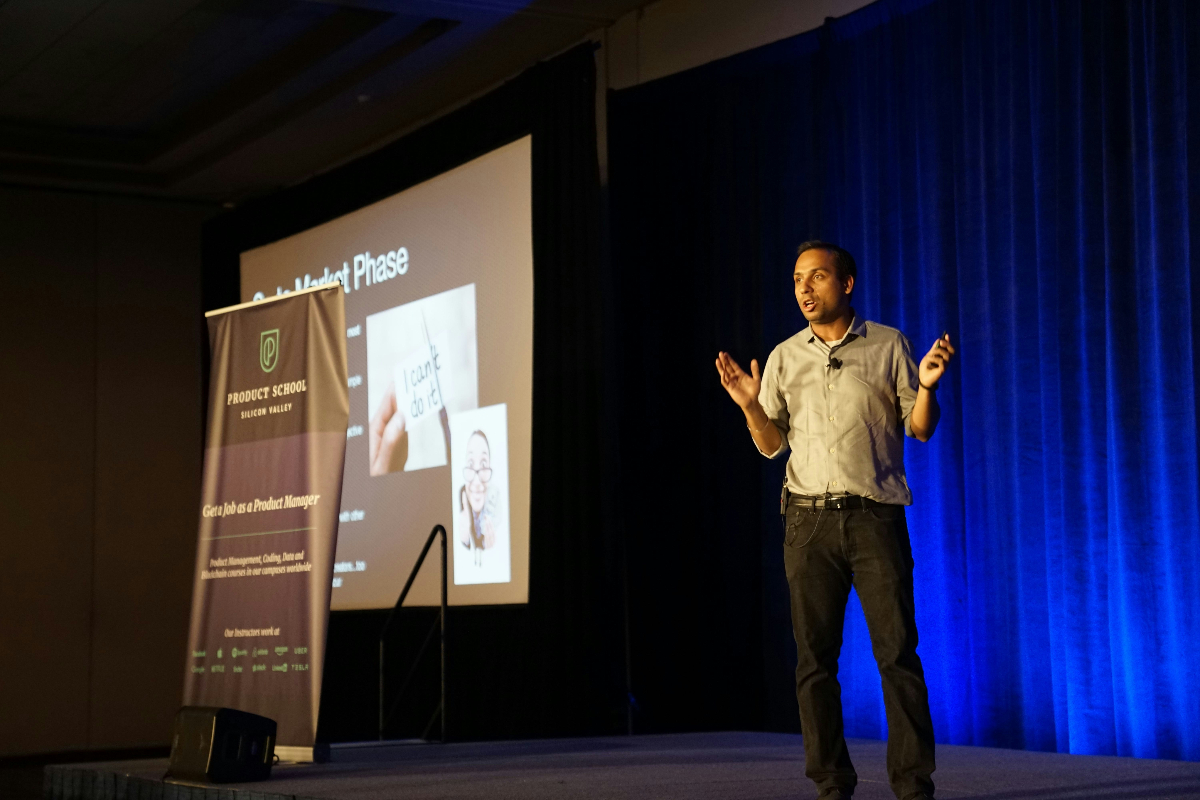 A man wearing a tan-colored shirt gestures on stage during sales pitch certification